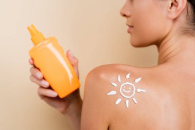 partial view of woman with image of happy sun on shoulder holding bottle of sunblock isolated on beige clipart