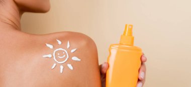 partial view of woman with bottle of sunblock and image of happy sun on naked shoulder isolated on beige, banner clipart