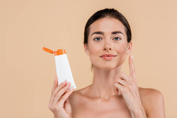 brunette woman holding sunscreen and pointing up with finger isolated on beige
