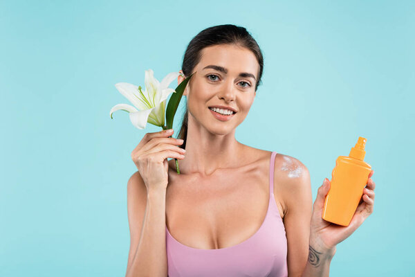 cheerful woman with sunblock and white lily looking at camera isolated on blue