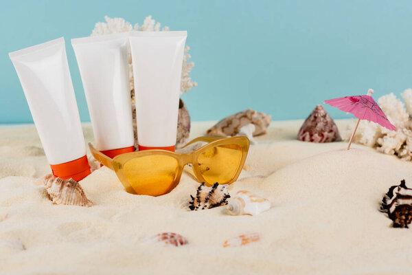 tubes of sunblock near orange sunglasses and seashells on sand isolated on blue