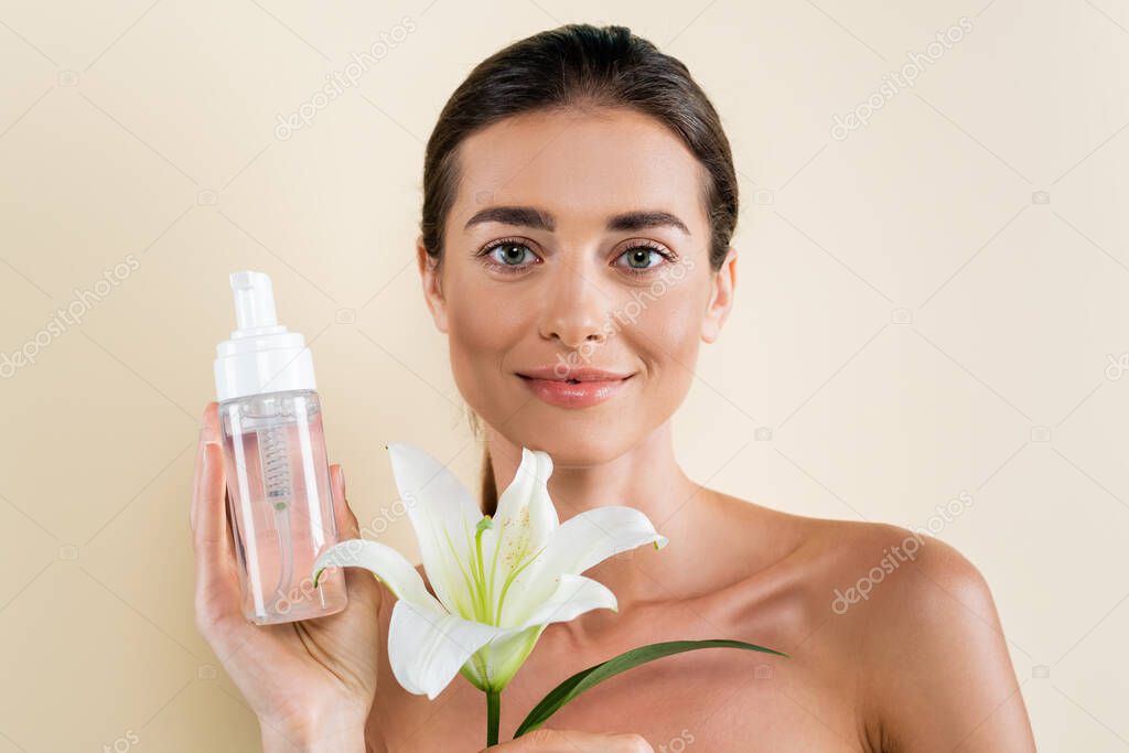 pleased woman with white lily and face tonic looking at camera isolated on light beige