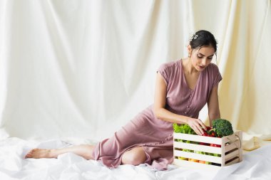 Esmer kadın kırmızı bibere ulaşıyor, sebzelerin yanında, beyaz kaptaki ahşap kutuda.