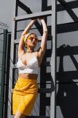 hezká žena v slunečních brýlích a žlutá šála pózující v blízkosti žebříku a betonové zdi 