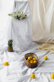 ripe lemons in wicker basket near pineapple on white bed sheets near bouquet of flowers 