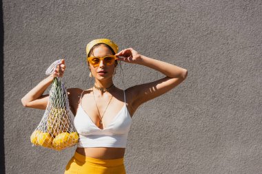 Güneş gözlüğünü ayarlayan, limon ve ananas dolu iple beton duvarın yanında duran şık bir kadın.