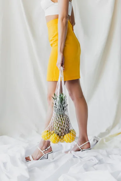 partial view of woman in yellow skirt and crop top holding string bag with lemons and pineapple on white