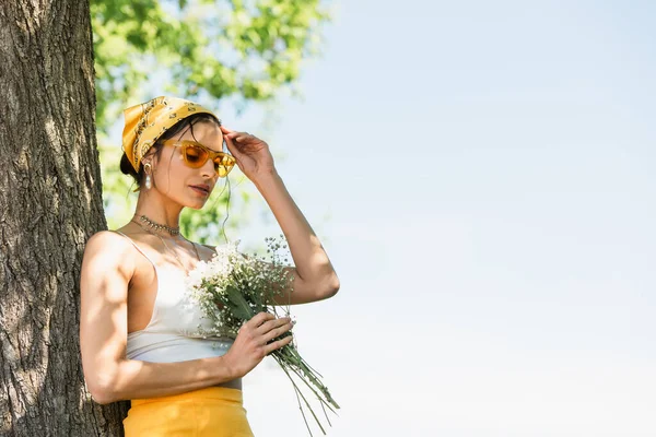 サングラスと黄色のスカーフの流行の女性は花を保持し 木の幹の近くに立って — ストック写真