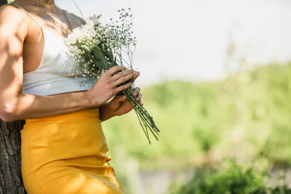 cropped view of woman in crop top holding flowers and standing near tree trunk