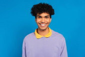 radostný africký Američan v šeříkovém svetru se žlutým límečkem usmívající se na kameru izolované na modré