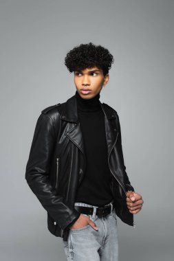 african american man in black leather jacket looking away while standing with hand in pocket of jeans isolated on grey