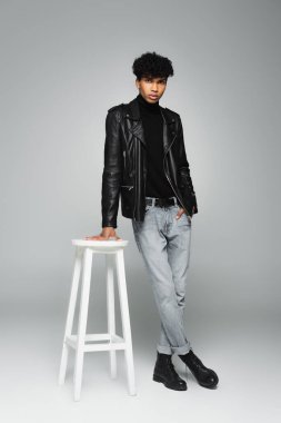 full length view of african american man in black leather jacket standing near white stool with hand in pocket on grey 