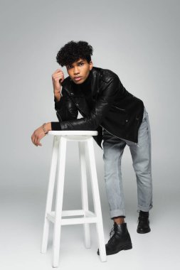 young african american man in black leather jacket and jeans leaning on high stool on grey background