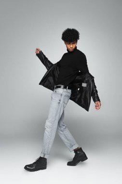 african american man in jeans, black turtleneck and leather jacket posing on grey 