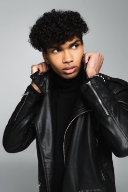 brunette african american man adjusting collar of black leather jacket isolated on grey