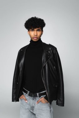 african american man with black leather jacket on shoulders standing with hands in pockets isolated on grey
