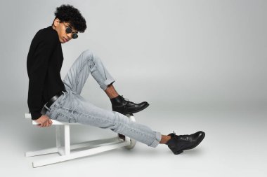 trendy african american man in black turtleneck, jeans and dark sunglasses sitting on overturned stool on grey