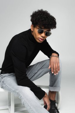 african american man in jeans, black turtleneck and dark sunglasses sitting on overturned stool on grey 