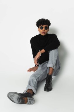young african american man in jeans and black turtleneck looking at camera while sitting on white 