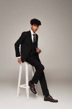 full length view of african american man in elegant formal wear sitting on high stool on grey background
