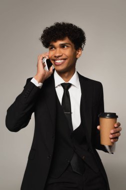 elegant african american man with coffee to go smiling and talking on smartphone isolated on grey clipart