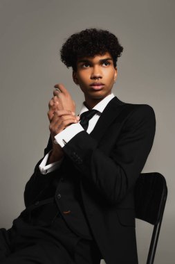 african american man in elegant three-piece suit looking away while sitting isolated on grey