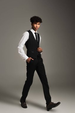 elegant african american businessman looking at camera while walking with hand in pocket on grey