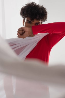 african american man looking at camera while obscuring face with arm near white blurred drapery