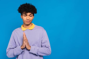  shamed african american man standing with praying hands gesture isolated on blue clipart