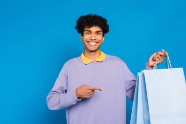 cheerful african american man pointing at shopping bags isolated on blue