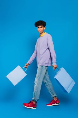 full length view of positive african american man walking with shopping bags on blue background
