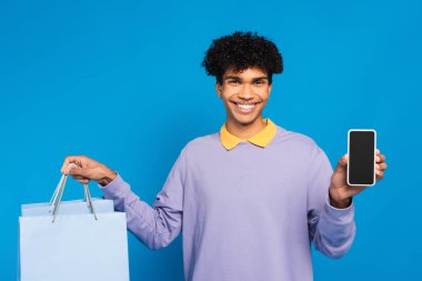 positive african american man with shopping bags and smartphone smiling at camera isolated on blue