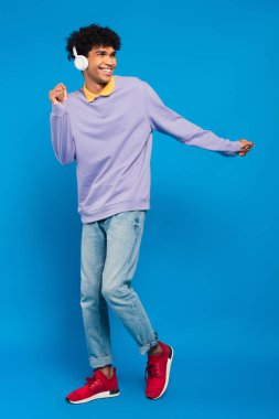 full length view of happy african american man dancing in headphones on blue background