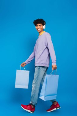 smiling african american man in headphones walking with shopping bags on blue background