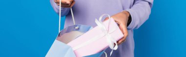 partial view of african american man putting gift into paper bag isolated on blue, banner