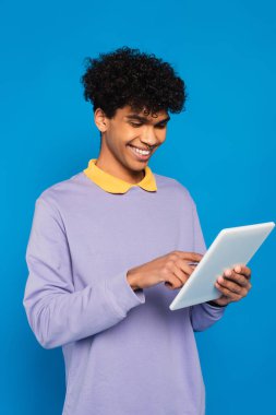 smiling african american man in violet jumper with yellow collar using digital tablet isolated on blue