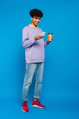 full length view of happy african american man with paper cup messaging on smartphone on blue background