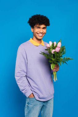 joyful african american man with bouquet holding hand in pocket isolated on blue