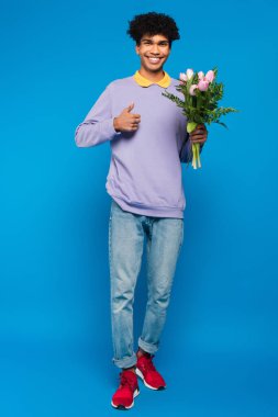 pleased african american man with bouquet of tulips showing thumb up on blue background