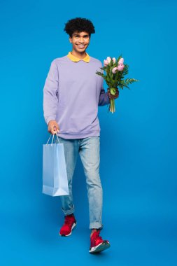 full length view of happy african american man walking with bouquet and shopping bag on blue background