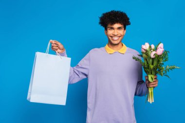 cheerful african american man showing flowers and shopping bag isolated on blue