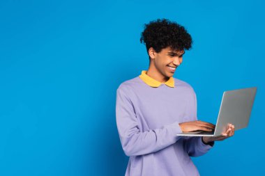 happy african american man in violet pullover using laptop isolated on blue