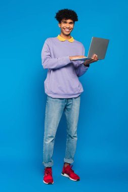 full length view of african american student using laptop on blue background