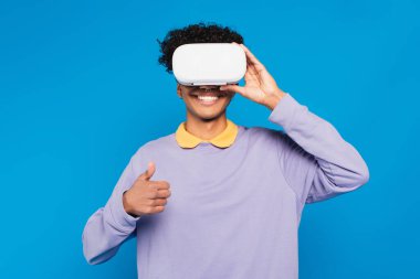 happy african american man in lilac jumper and vr headset showing thumb up isolated on blue