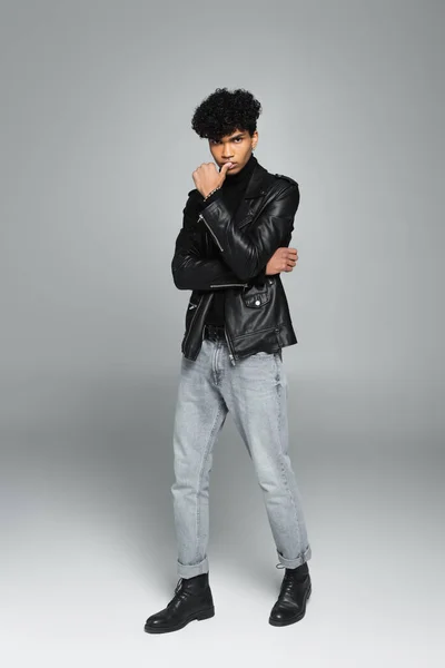 african american man in jeans, black leather jacket and boots posing with hand near mouth on grey background