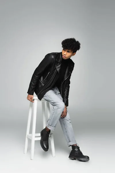 穿着牛仔裤和皮夹克的时髦的非洲裔美国人 在灰色背景的高凳子上远眺 — 图库照片