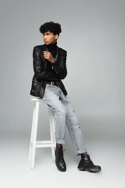 young african american man in leather jacket and jeans looking away on high stool on grey background