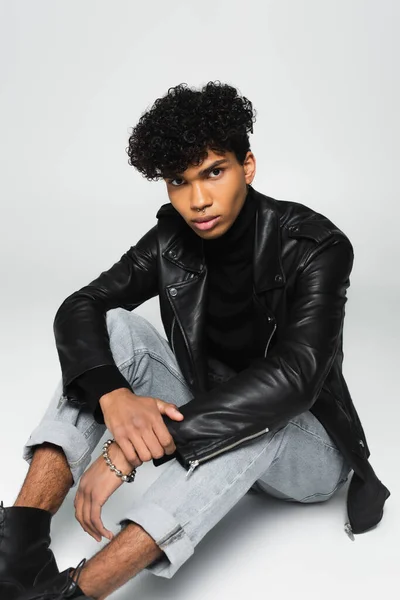 curly african american man in black leather jacket and jeans sitting on grey background