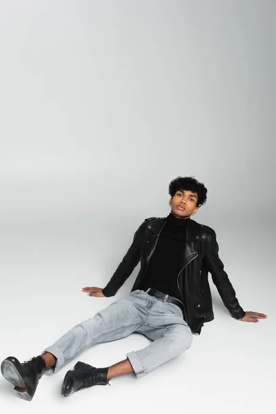trendy african american man in jeans and black leather jacket looking at camera while sitting on grey