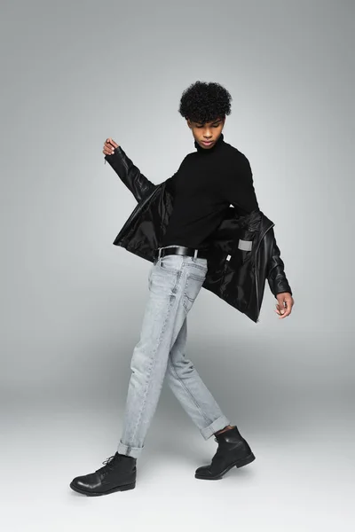 african american man in jeans, black turtleneck and leather jacket posing on grey